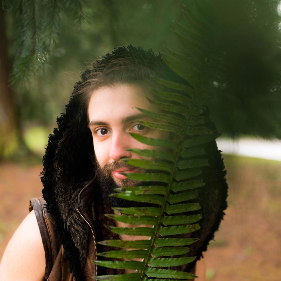 Bizio in a vest with a hood smiling behind a large fern